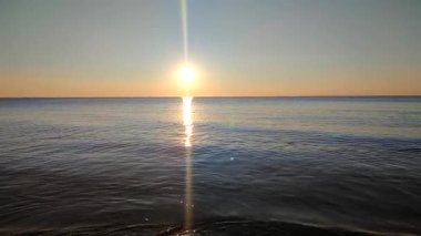Denizde gün batımı. Güneş ufukta görünüyor. Sakin mavi su yüzeyi, küçük dalgalar, parlayan güneş berrak bulutsuz gökyüzü, ufuk çizgisi, deniz yüzeyindeki güneş yolu, güneş ışınları. Ağır çekim. Güzel doğal arkaplan