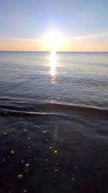 Denizde gün batımı. Güneş ufukta görünüyor. Sakin mavi su yüzeyi, küçük dalgalar, parlayan güneş, bulutsuz gökyüzü, ufuk çizgisi, deniz yüzeyindeki güneş yolu, güneş ışınları. Ağır çekim. Doğal arka plan. Dikey video