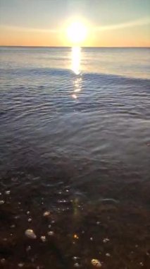 Denizde gün batımı. Güneş ufukta görünüyor. Sakin mavi su yüzeyi, küçük dalgalar, parlayan güneş, bulutsuz gökyüzü, ufuk çizgisi, deniz yüzeyindeki güneş yolu, güneş ışınları. Ağır çekim. Doğal arka plan. Dikey video