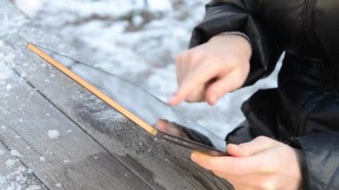 Tablette çalışan bir kız bir kış günü açık havada, sokağın yakınındaki tahta bir masada oturuyor. Beyaz ırk