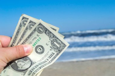 Elinde beyaz köpüklü, kumlu ve güneşli yaz gününde açık mavi gökyüzü olan deniz ve deniz dalgalarının arka planında üç dolarlık banknot tutan kişi. Para kavramı, tatil masrafları, seyahat, turizm