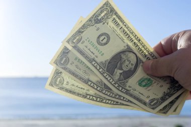 Elinde bir dolarlık banknotlar tutan bir adam denizin arka planına, kumlu kumsala ve güneşli yaz gününde mavi gökyüzüne karşı. Para, finans, para, seyahat, turizm, tatil, dinlenme kavramı