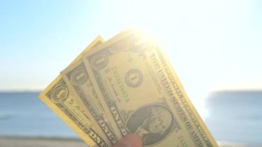 Elinde dolar banknotları tutan kişi. Parlak güneş, mavi gökyüzü ve durgun mavi denizin arka planında bir dolar değerinde. Bir dolarlık banknotlar mavi deniz gökyüzünün zemininde