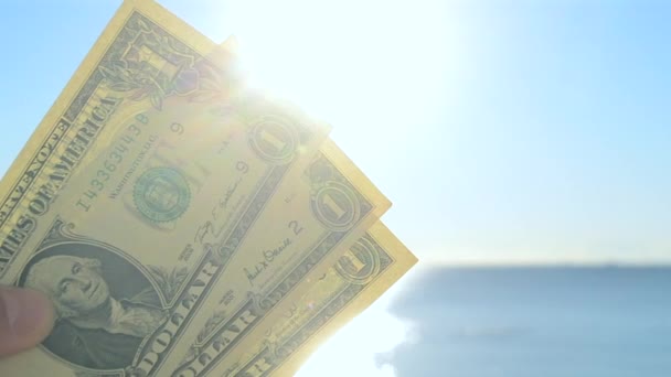 明るく輝く太陽 青い空と穏やかな青い海を背景に1ドルの顔の値を持つドルの手札を保持する人 青い空を背景に1ドル紙幣 — ストック動画