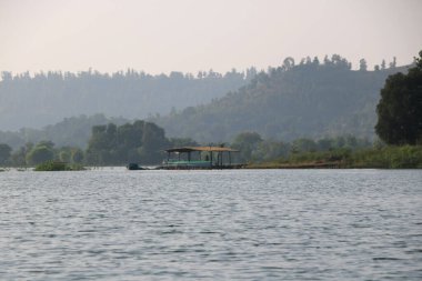 Nehir kıyısına yakın küçük kulübe ve tekne parkı manzarası. Arka planda tepeler ve puslu mavi gökyüzü.