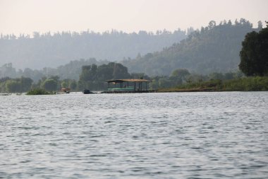 Nehir kıyısına yakın küçük kulübe ve tekne parkı manzarası. Arka planda tepeler ve puslu mavi gökyüzü.