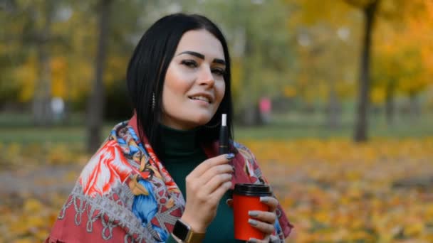 Portret pięknej brunetki w jesiennym parku pije kawę i pali elektronicznego papierosa — Wideo stockowe