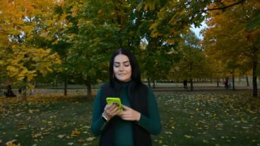 Sonbahar parkında gülümseyen ve mesaj yazan güzel bir genç kadın.