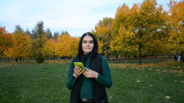 Sonbahar parkında gülümseyen ve mesaj yazan güzel bir genç kadın.