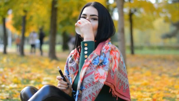 Красива стильна молода дівчина портрет брюнетка п'є гарячу каву і курить електронну сигарету в осінньому парку — стокове відео