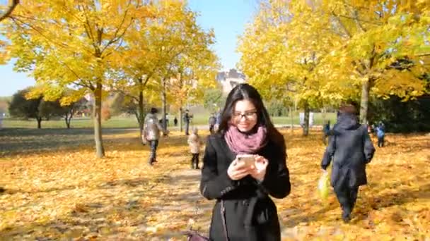 Una chica morena en gafas camina en un parque otoñal sonriendo y escribe un mensaje — Vídeo de stock