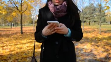 Sonbahar parkında yürüyen ve akıllı telefon kullanan mutlu bir kadının ön görüntüsü.