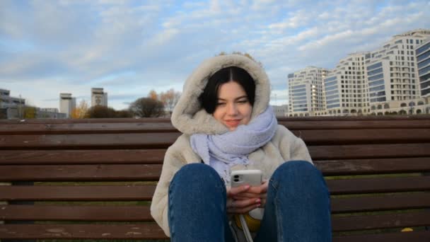 Fiatal csinos nő mobiltelefon ül padon az őszi parkban, és görgetés a szociális hálózat — Stock videók