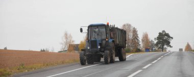 Sonbahar günü, arka planda sarı huş ağaçları üzerinde asfalt yolda römorklu bir traktör, Avrupa tarım ulaşım makineleri