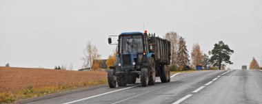 Mavi tekerlekli traktör boş asfaltlanmış yol kenarında sonbahar günü ağaçların arkasında Rus tarım lojistiği.
