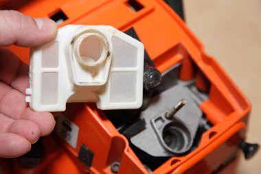 Bir adamın elindeki elektrikli testere hava filtresi karbüratör arka planının kapağının altında, kullanımdan sonra bahçe elektrikli aletlerini kontrol edip temizler