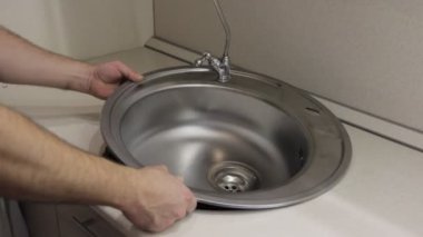 Yeni gri paslanmaz çelik metal kevgir lavabo tesisatı ve yumuşak boruları olan su musluğu tesisatçı beyaz elleriyle beyaz mutfak masasına koyuyor.