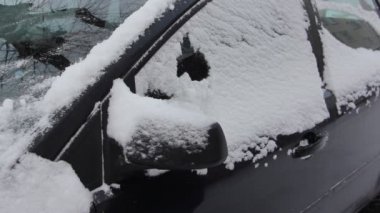 Kırmızı fırçalı kış eldiveni giymiş bir adam kar kaplı bir arabanın yan camını temizler.