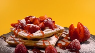 Çilekli Belçika waffleları, tatlı çilek soslu, krem şantili ve pudra şekerli turuncu arka planda bir taş tahtada.