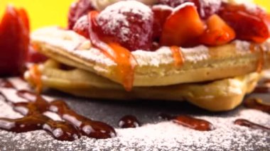 Çilekli Belçika waffleları, tatlı çilek soslu, krem şantili ve pudra şekerli turuncu arka planda bir taş tahtada.