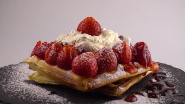 Çilekli Belçika waffleları, tatlı çilek soslu, krem şantili ve pudra şekerli beyaz arka planda bir taş tahtada.
