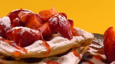 Çilekli Belçika waffleları, tatlı çilek soslu, krem şantili ve pudra şekerli turuncu arka planda bir taş tahtada.