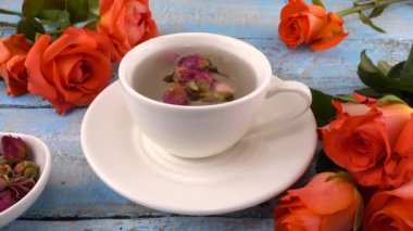 White cap of  tea with fragrant dried rose buds, fresh rose flowers on vintage light blue wooden tabletop. Tea brewing.