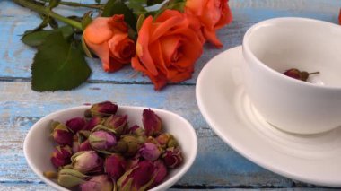 White cap of  tea with fragrant dried rose buds, fresh rose flowers on vintage light blue wooden tabletop. Tea brewing.