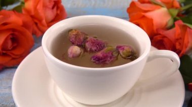 White cap of  tea with fragrant dried rose buds, fresh rose flowers on vintage light blue wooden tabletop. Tea brewing.