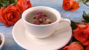 White cap of  tea with fragrant dried rose buds, fresh rose flowers on vintage light blue wooden tabletop. Tea brewing.