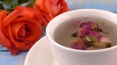 White cap of  tea with fragrant dried rose buds, fresh rose flowers on vintage light blue wooden tabletop. Tea brewing.