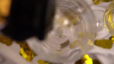 Bir şişe köpüklü şaraptan konfeti arkaplanındaki bardaklara dökülüyor. Yavaş çekim.