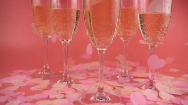 Pembe arka planda köpüklü şarap kadehleri ve kalp şeklinde konfetiler. Yavaş çekim.