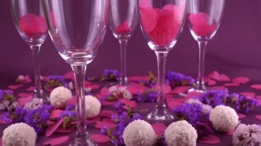Sparkling wine is poured into a glass on a purple background with confetti, flowers and round candy with coconut flakes. Falling confetti in the form of hearts. Slow motion.