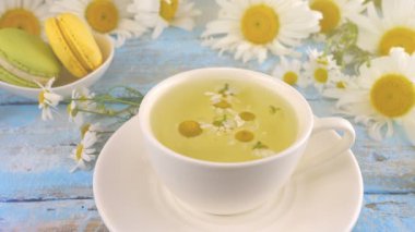 Papatya çiçekleri, bitki çayı ve klasik açık mavi ahşap masa üstünde badem kurabiyesi kompozisyonu. Papatya çayını beyaz fincanda demliyorum. Yaz teması.