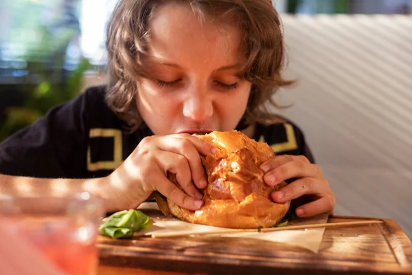 happy boy bites hot hamburger with pork cutlet, lettuce, tomato, pickle in interior. Summer day