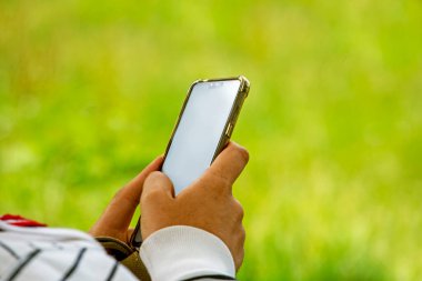 Adam elinde piknikte çimlerin üzerinde uzanmış beyaz bir akıllı telefon tutuyor. Reklam için Smartphone şablonu kullan.