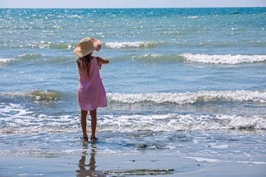 happy little girl in a hat with her back is played with water on the sea surf. Family holiday