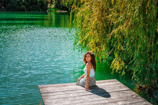一位穿着裙子的迷人的年轻女子正坐在一座木制的桥上 面对着森林中的一个湖泊的大自然 积极生活方式的概念 — 图库照片