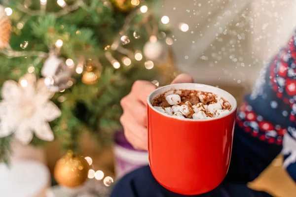 A man holds a mug of cocoa with marshmallows in front of a Christmas tree