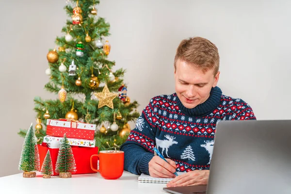 A young man in a sweater signs postcards and writes letters next to a Christmas tree. The concept of preparation for the celebration of New Year and Christmas
