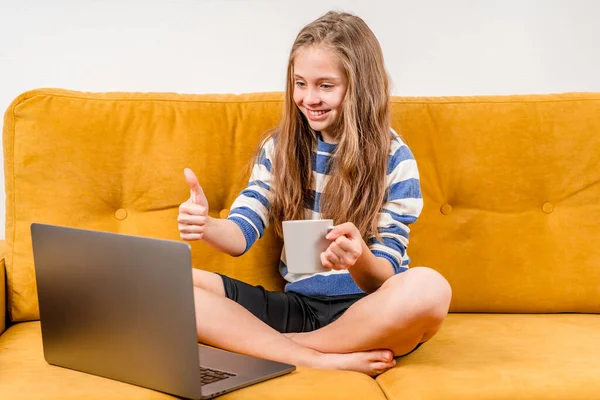 笑顔ヨーロッパの10代の女の子は黄色のソファに座っている友人や両親へのビデオ通話のためのラップトップを使用しています 遠隔オンライン教育 — ストック写真