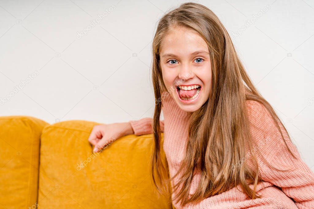 Beautiful little girl makes a face and fooling around sitting on a yellow sofa