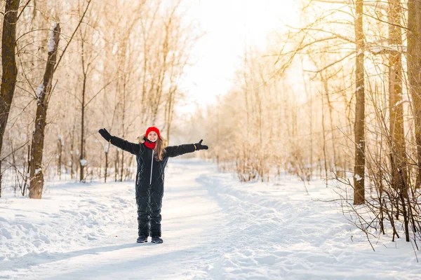 冬の森の中で日光のある赤い帽子の少女10歳 小さな女の子は冬の日に楽しんでいます 暖かいジャンプスーツを着た陽気な女の子 — ストック写真