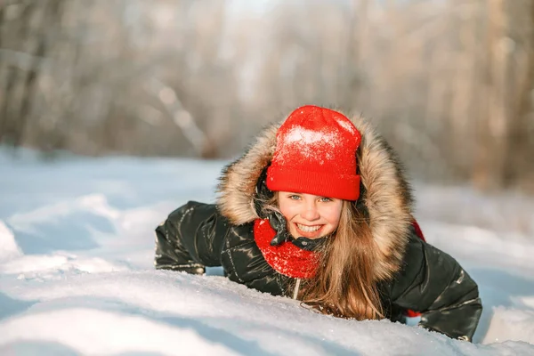 ピンクの頬をした幸せな女の子が雪のドリフトに横たわっていて 美しい冬の森の中で笑う — ストック写真