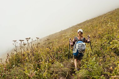 Ekose gömlekli, büyük sırt çantalı, sis içinde dağ yolunda yürüyen, sağlıklı aktif yaşam tarzı, dijital detoks, hafta sonu aktiviteleri olan genç bir kadın.