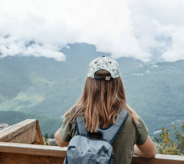 Rear view of young blonde woman in cap with backpack looking at view of Caucasian mountains and cloudy sky standing on wooden viewpoint, hiking active healthy lifestyle enjoying nature