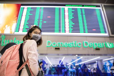 Maskeli bir kadın, pembe bir sırt çantasıyla havaalanının uçuş takvimine bakıyor.