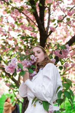 Beyaz gömlekli sarışın bir kadın kiraz çiçeklerinin yanında poz veriyor. Bahar havası