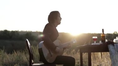 Müzisyen gün batımında bir tarlada gitar çalar. Şarap kadehli ahşap masa. Kırsal alan.